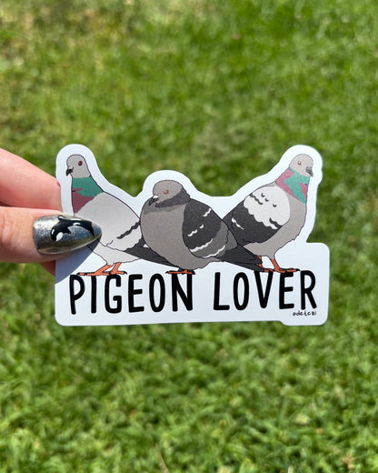 A hand with silver nail polish holds a die-cut sticker featuring four pigeons and the text 'PIGEON LOVER' against a blurred green grass background.