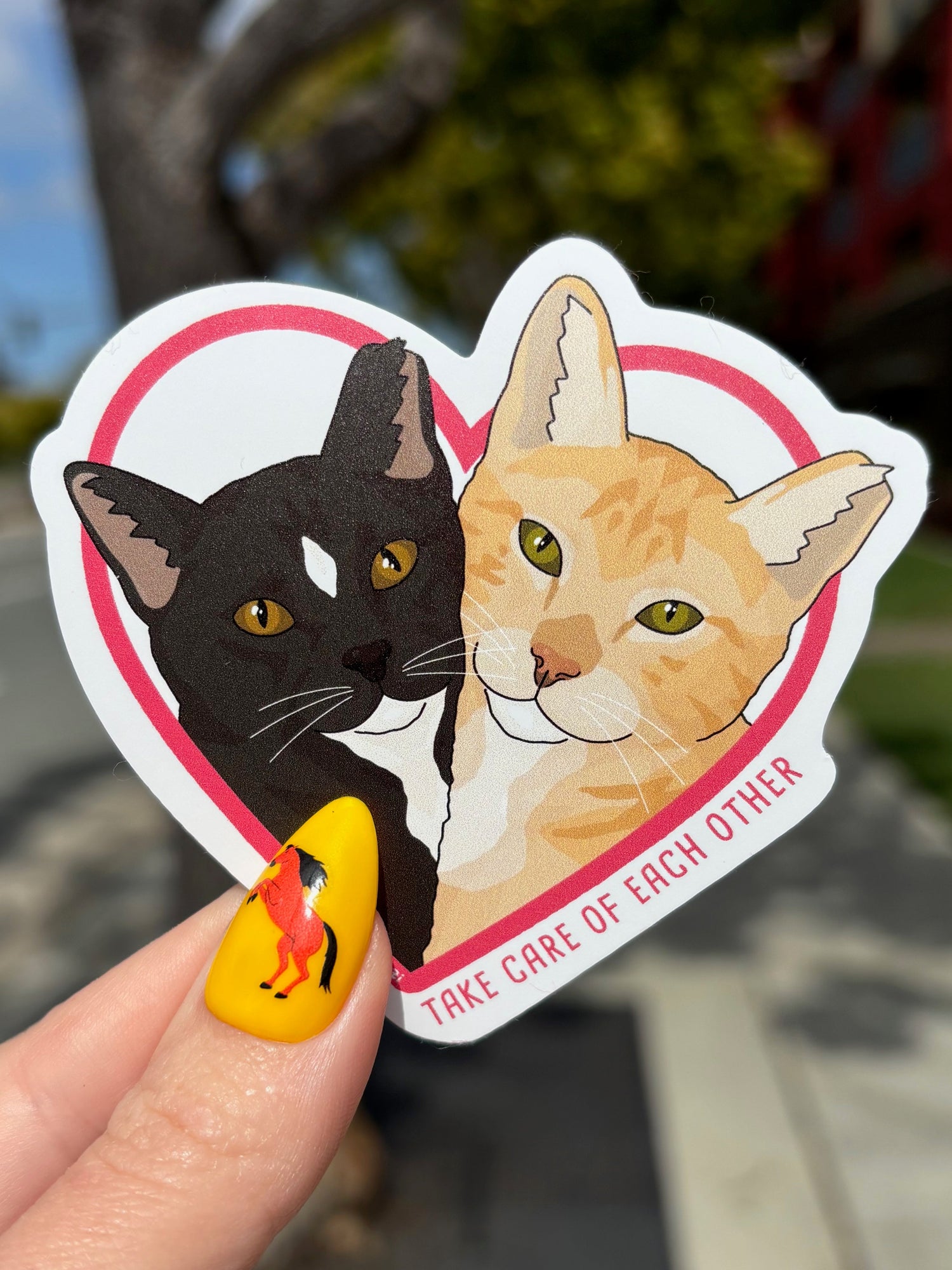 Heart-shaped sticker with two cats and text held by a hand outdoors.